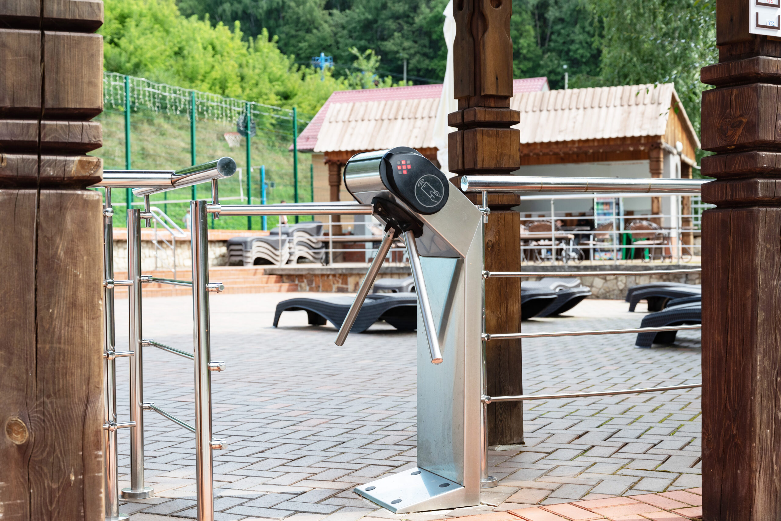 Turnstile gate at entry of an outdoor swimming pool, surrounded by wooden pillars and metal railings, with greenery in the background, concept of access control