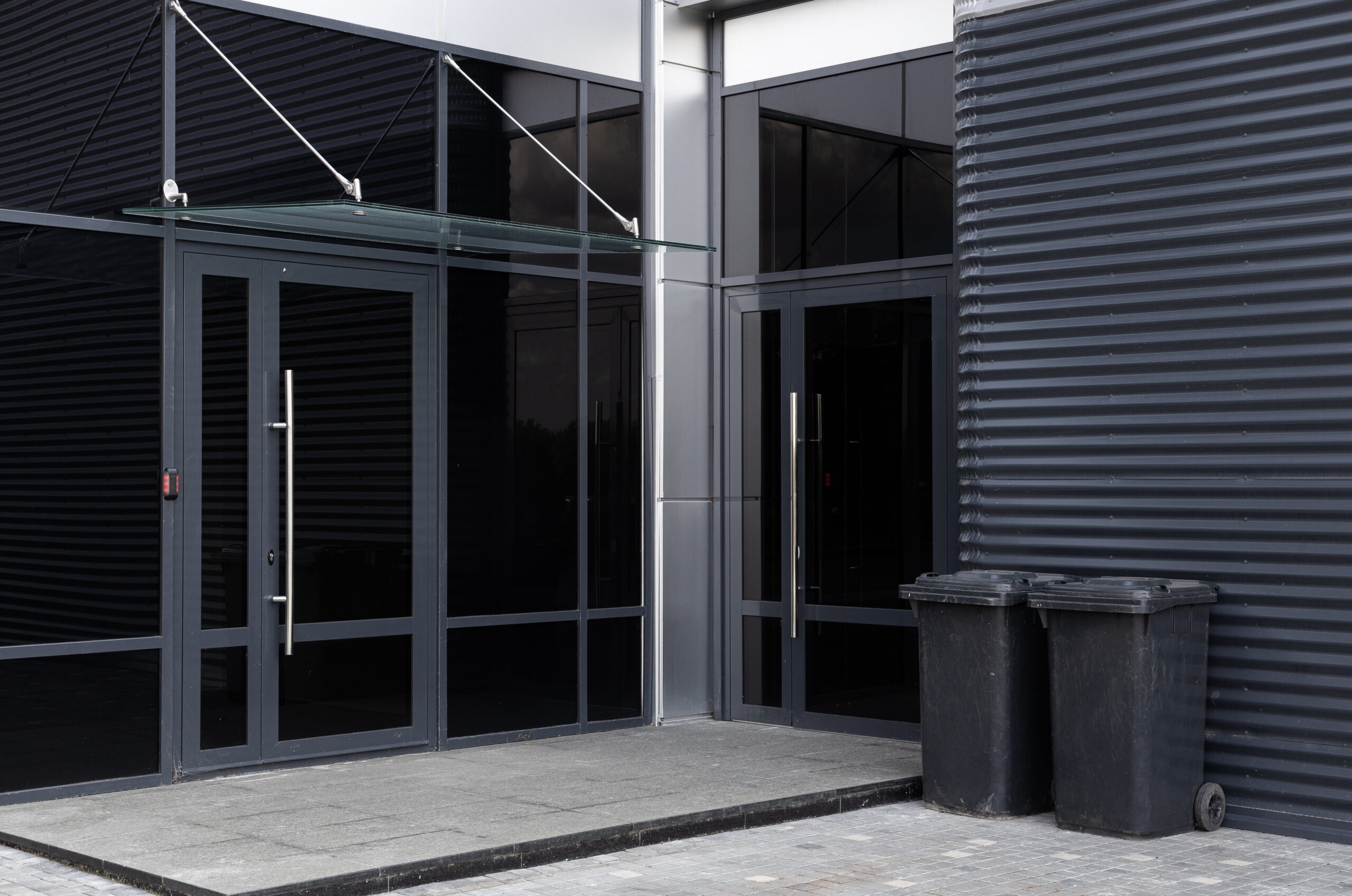 Glass doors - entrance to modern building and two scuffed black dustbins against black metal corrugated wall nearby